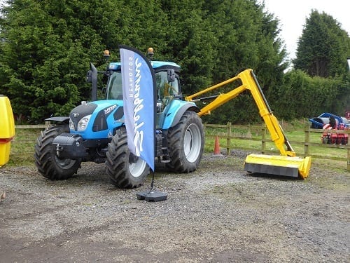 A blue tractor equipped with Shelbourne Reynold Hedge Cutters is parked in front of a fence.